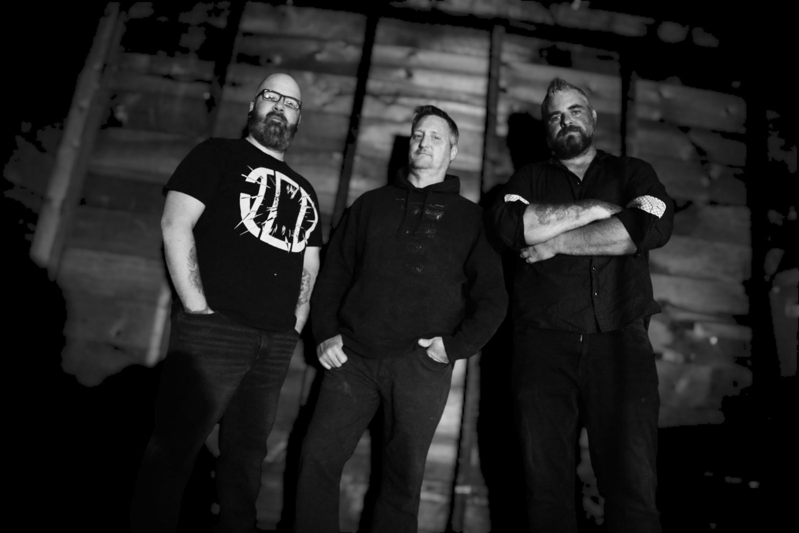 Three members of Undercard, Josh Eberhard, Alex Vota, Mike Forland from left to right, standing in front of a wooden backdrop, dressed in black, with serious expressions, capturing the band's dark aesthetic.