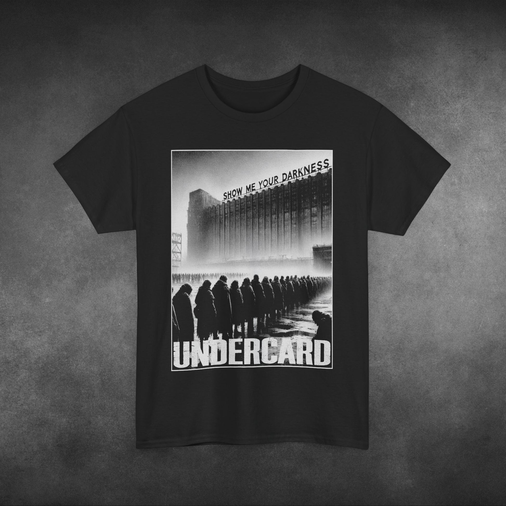  A black t-shirt featuring a monochrome, dystopian-style graphic of people standing in line in front of a large, industrial building with the text "SHOW ME YOUR DARKNESS" above. The word "Undercard" is boldly printed at the bottom of the design in distressed white lettering. The image conveys a dark, moody aesthetic, with the t-shirt set against a textured grey background.